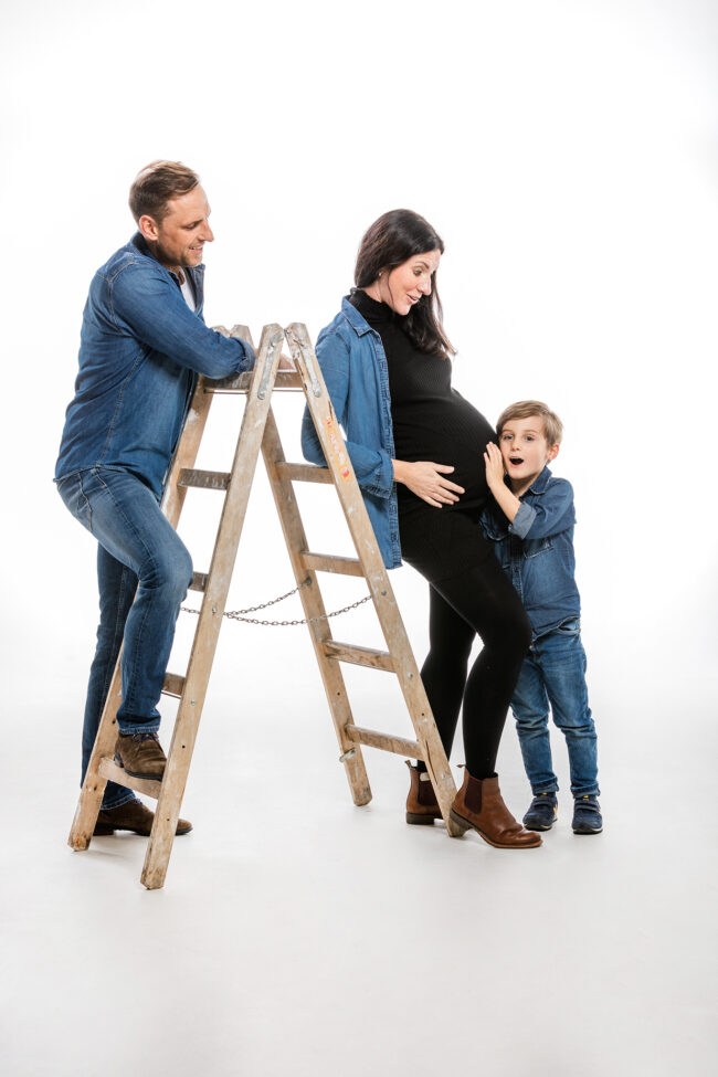 Familie, Vater und Mutter mit Babybauch lehnt an Leiter, Kind lauscht am Bauch, Studioaufnahme vor weißem Hintergrund