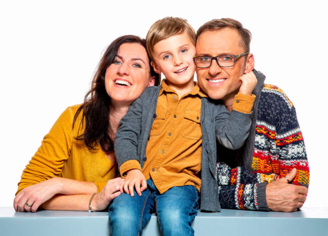 Familie, Eltern mit Kleinkind, Kind sitz auf Bank, Eltern knien dahinter, alle lachen in die Kamera