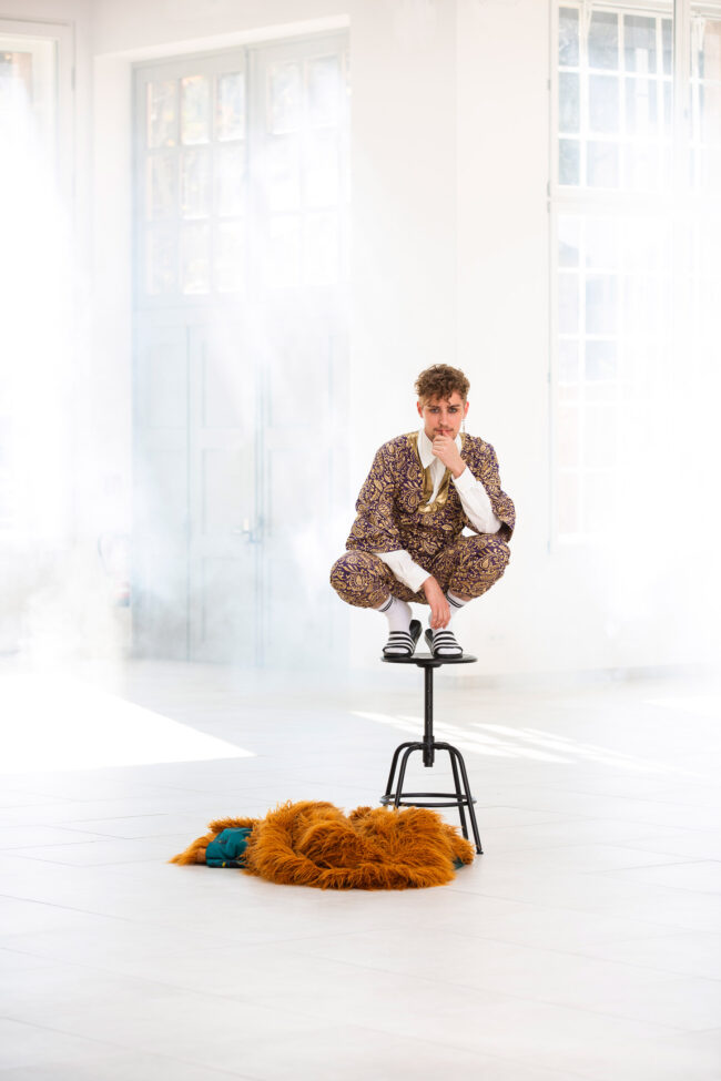 Fashion, männliches Model in gold-violettem gemustertem Einteiler, weißen Tennissocken und Adiletten auf schwarzem Stuhl hockend, hellbrauner Fellmantel am Boden liegend, vor hellem nebeligem Hintergrund mit Fenstern