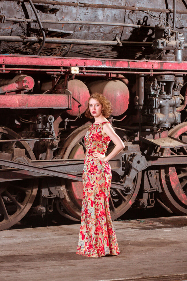 Fashion, Model in besticktem Oberteil steht vor einer alten Lokomotive, industrieller Hintergrund, Hochformat
