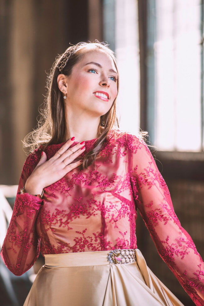 Fashion, Model in dunkelrosanem bestickten Oberteil und hellrosanem Rock, Hand an den Hals gelegt, Halbaufnahme, vor unscharfem Fenster im Hintergrund