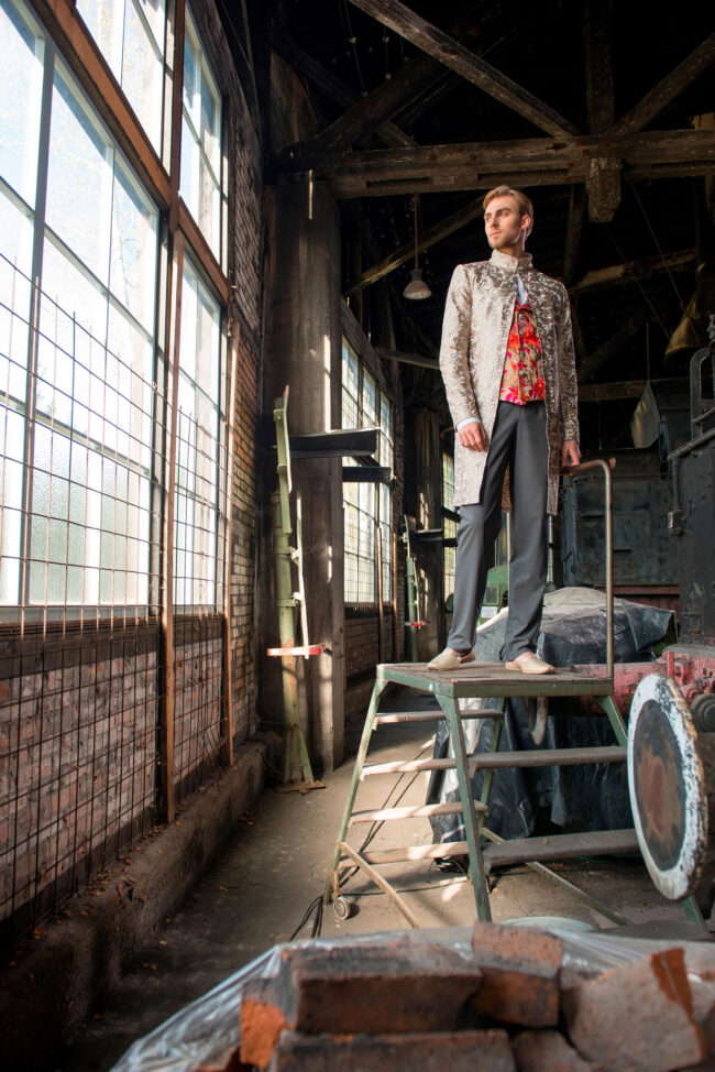 Fashion, Model in besticktem Oberteil und silbernem Mantel auf einer Treppe, vor industriellem Hintergrund, Hochformat