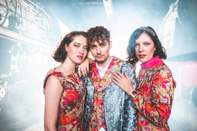 Fashion, 3 Models in bestickten Oberteilen vor industriellem Hintergrund, Beleuchtung von hinten, Querformat