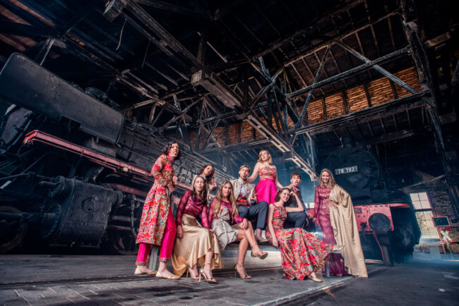 Fashion, Modelgruppe in bestickten Kleidern vor industriellem Hintergrund mit alter Dampflokomotive, Weitwinkel, Querformat