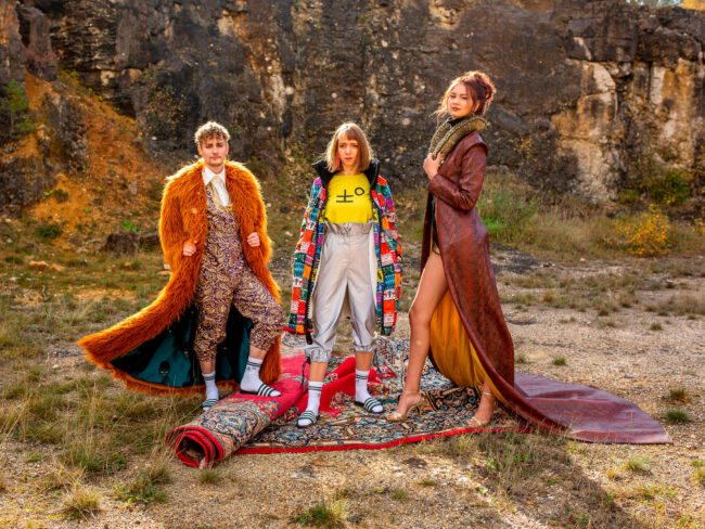 Fashion, 3 Models in verschiedenen bunten Outfits stehen auf Teppich vor Felswand, schauen in die Kamera, Querformat, Ganzaufnahme