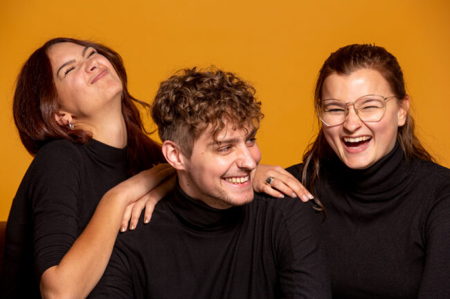 Freundschaftsshooting, drei Freunde vor gelbem Hintergrund lachen in die Kamera