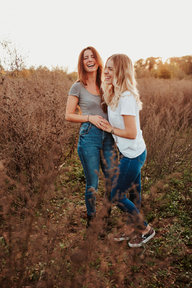 Freundschaftsshooting, zwei Frauen in getrocknetem Blumenfeld. lachen in die Kamera und halten sich an den Händen, Ganzaufnahme