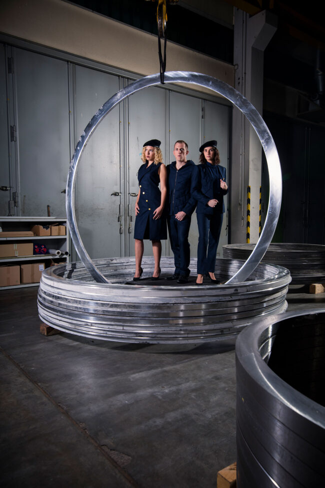 Fashion, 3 Models in dunkelblauen Kostümen und Barrett in Metallringen stehend vor industriellem Hintergrund, Hochformat, Ganzaufnahme