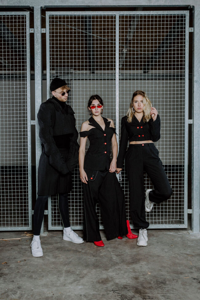 3 schwarz gekleidete Models mit weißen/roten Schuhen lehnen an/stehen vor einem Metallgitter Tor