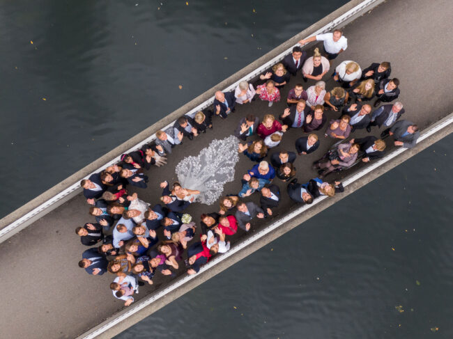 Hochzeit, Gruppenaufnahme, Brautpaar und Gäste auf einer Brücke die nach oben winken, Drohnenaufnahme, Querformat