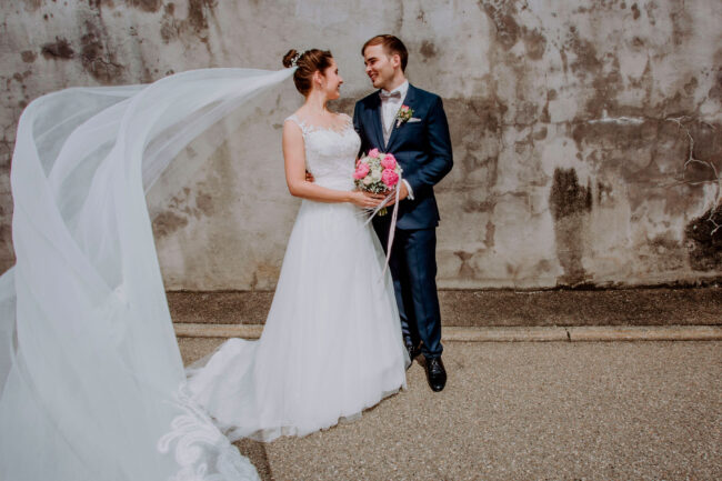 Hochzeit, Portrait, Paar hält sich im Arm und lacht sich an, Schleier wird zur seite geweht, Querformat