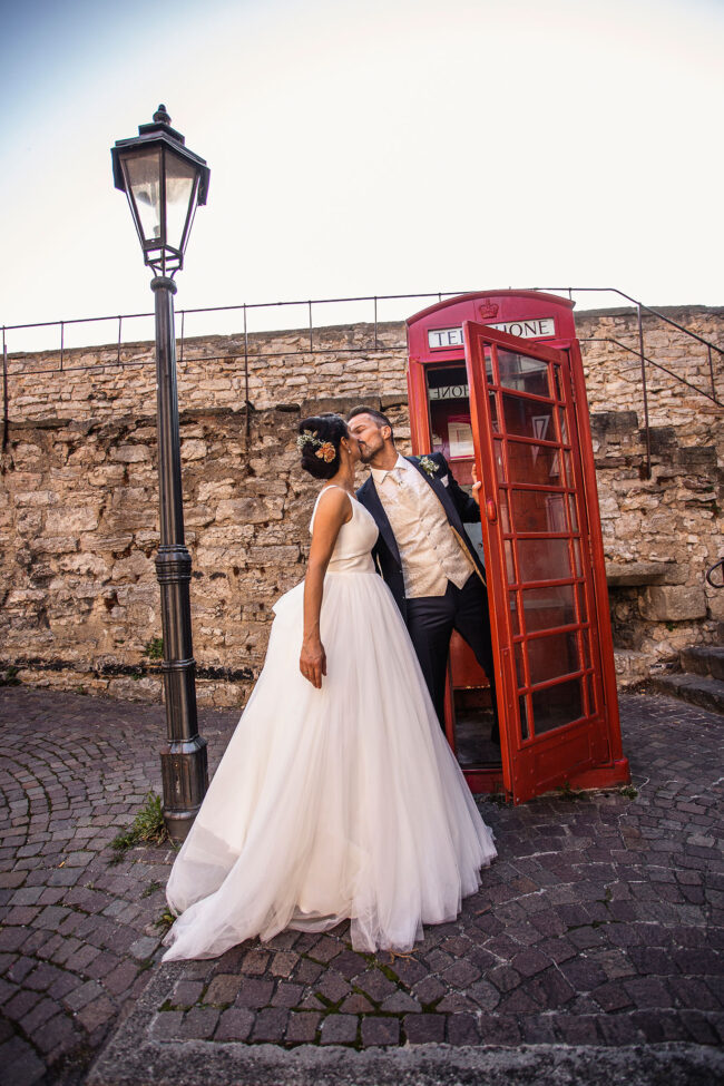 Hochzeit, Bräutigam lehnt sich aus roter Telefonzelle und küsst die Braut