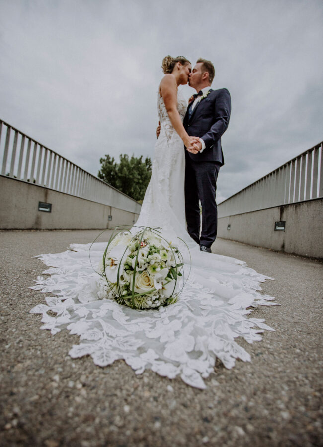 Hochzeit, Portrait, Paar hält sich im Arm und küsst sich, Brautstrauß liegt auf Schleppe, Paar steht auf einer Betonbrücke