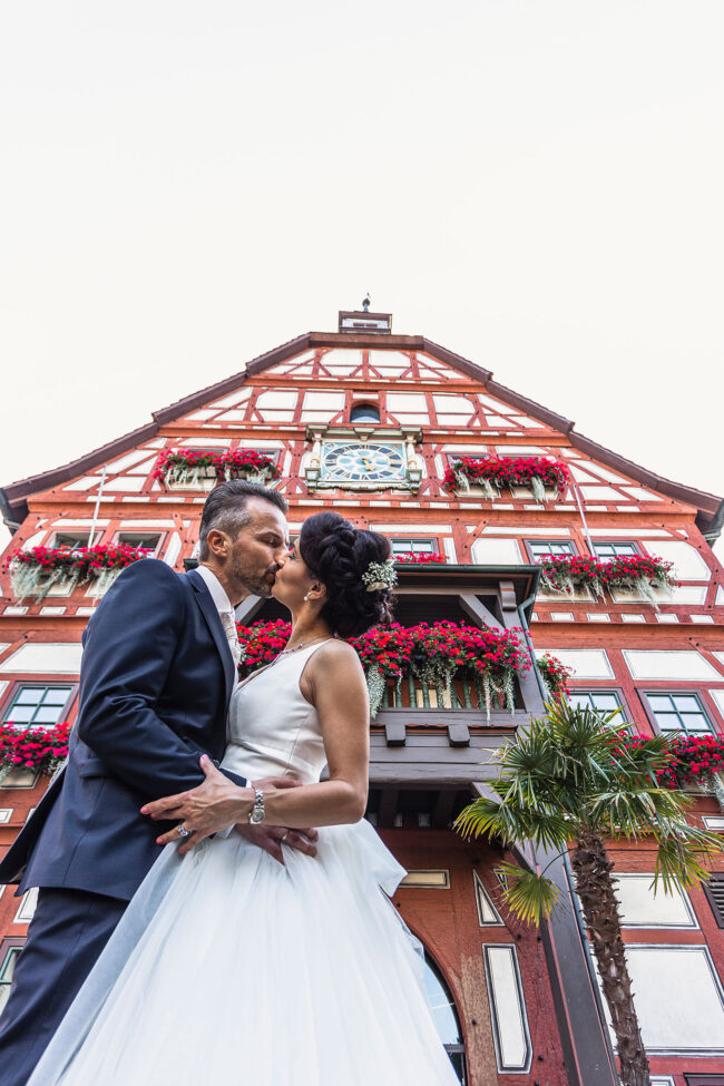 Hochzeit, Portrait, Halbaufnahme von küssendem Paar von unten vor einem roten Fachwerkhaus mit Blumen an den Fenstern
