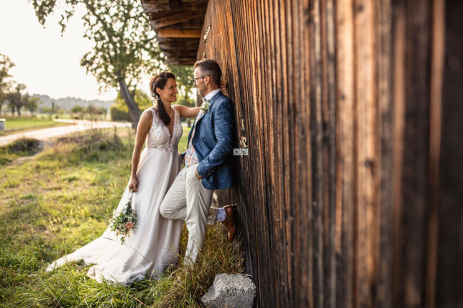 Hochzeit, Portrait, Bräutigam steht lässig an eine Holzwand gelehnt, Braut steht vor Mann, beide schauen sich an