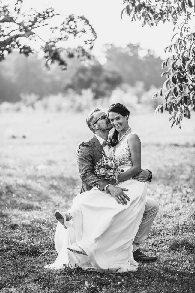 Hochzeit, Portrait, Bräutigam sitze auf einer Wiese und hat Braut auf dem Schoß. Er gibt ihr einen Kuss auf die Wange, sie lächelt und steckt die Beine in die Höhe, Hochformat, schwarz-weiß