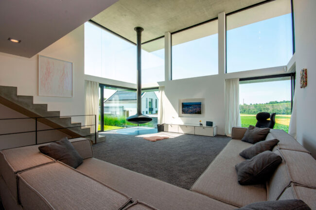 Interieur Wohnbereich mit weitläufigem grauem Sofa, im Hintergrund freihängender Kamin vor bodentiefer Fensterfront mit Aussicht auf Weinberge