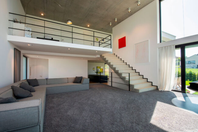 Innenaufnahme, Wohnbereich mit großem grauen Sofa, eine Treppe führt zu einer offenen Galerie im oberen Stockwerk