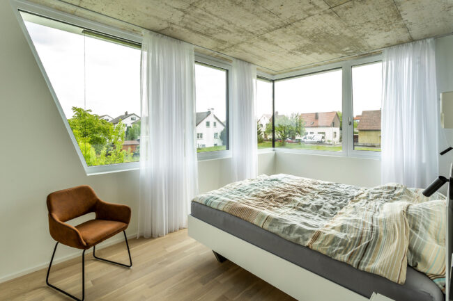 Interieur Schlafbereich mit Bett, Fenster mit weißen Vorhängen, heller Holzboden, ein brauner Sessel steht an einer Wand