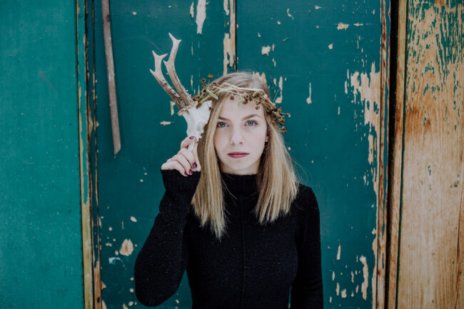 Portrait, Frau mit kleinem Hirschgeweih und Blumenkrone vor petrolfarbener Holzwand