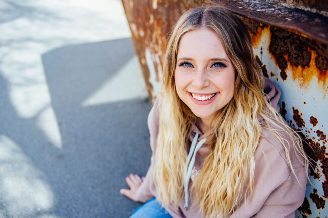 Portrait, Frau sitzt vor rostiger Wand und schaut lächelnd nach oben in die Kamera