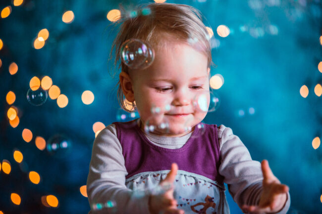Famili, Kind in lilanem Kleid fängt Seifenblasen, vor blauem Hintergrund