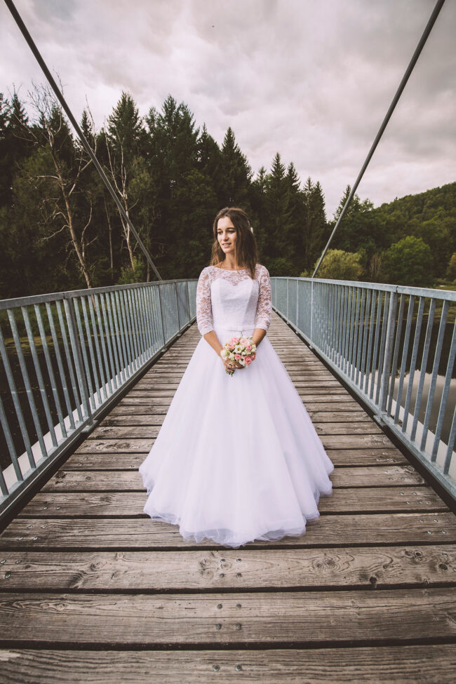 Hochzeit, Portrait, Braut steht auf Brücke und schaut in die Ferne, im Hintergrund ist Wald