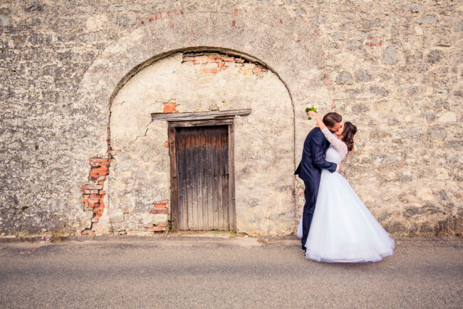 Hochzeit, Portrait, Brautpaar hält sich küssend im Arm und steht vor einer alten Steinwand mit Holztür in der Mitte
