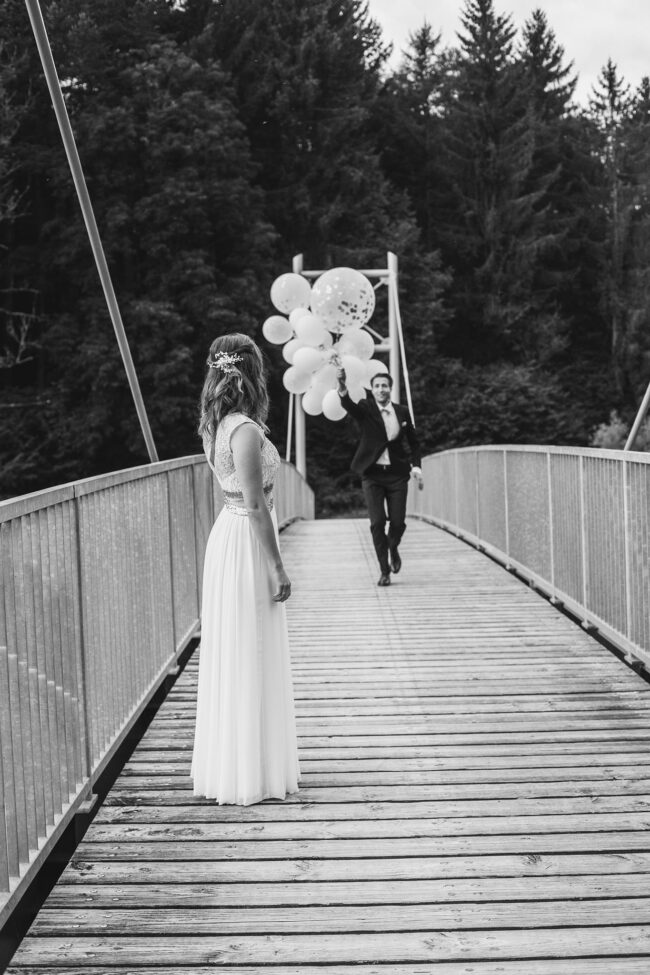 Hochzeit, Portrait, Braut steht auf Brücke und Bräutigam kommt mit weißen Luftballons auf sie zu, schwarz-weiß, Ganzaufnahme