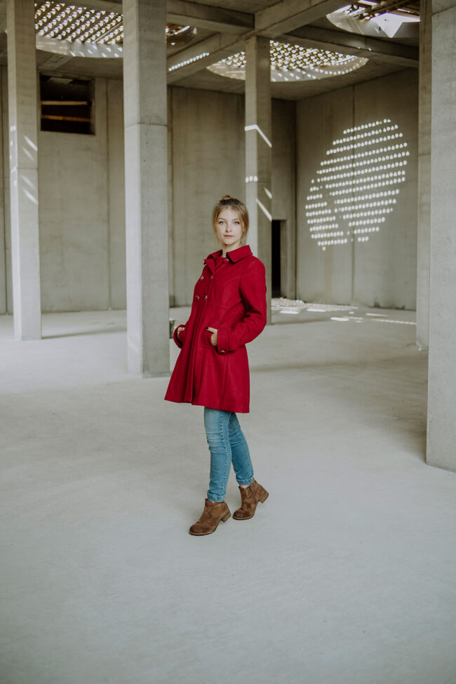 Fashion, Portrait, junge Frau in rotem Mantel und Betonhalle mit Lichtschächten, Ganzaufnahme