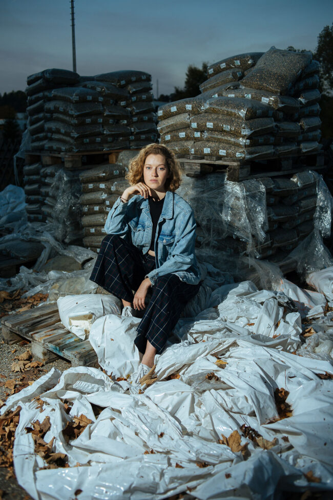 Portrait, junge Frau sitzt auf mit Plastik bedeckten Holzpaletten auf Wertstoffhof, Ganzaufnahme