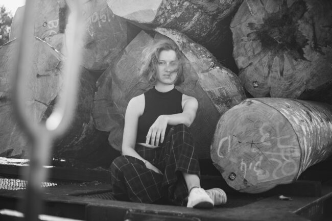 Portrait, Frau sitzt vor aufgeschichteten Baumstämmen, Schwarz-Weiß