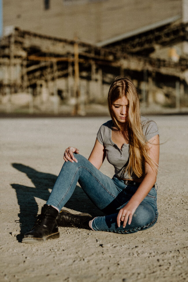 Portrait, junge Frau in Jeans und T-Shirt sitzt auf Boden vor Industriegelände, Ganzaufnahme