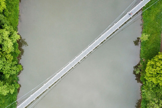 Drohnenaufnahme, Landschaft, weiße Hängebrücke über Fluß mit Gründer Uferböschung