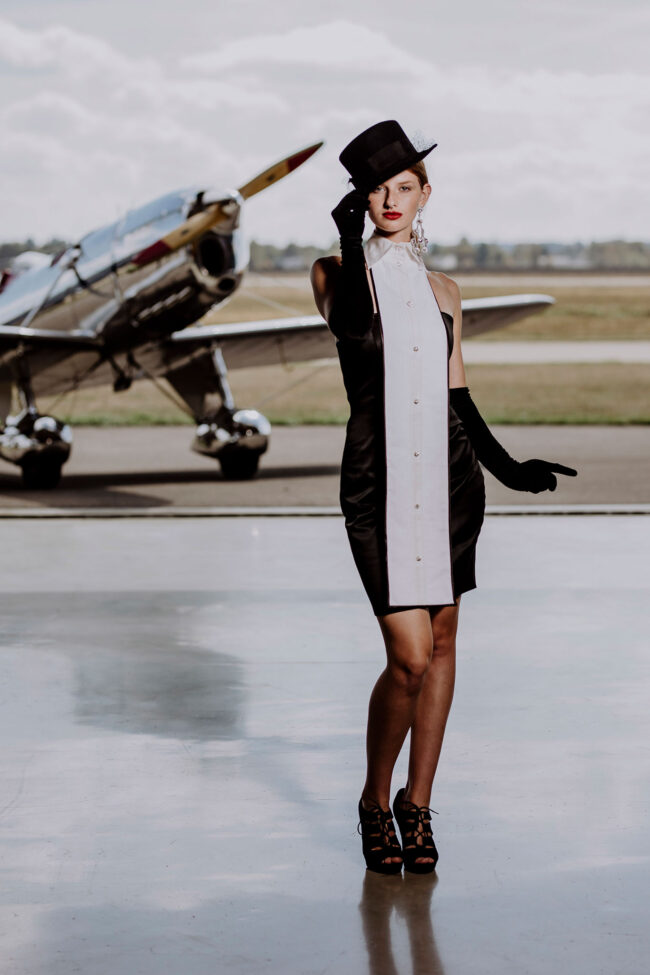 Fashion, Model mit schwarzem Hut, Handschuhen und weiß-schwarzes Kleid, vor Propellerflugzeug, Hochformat