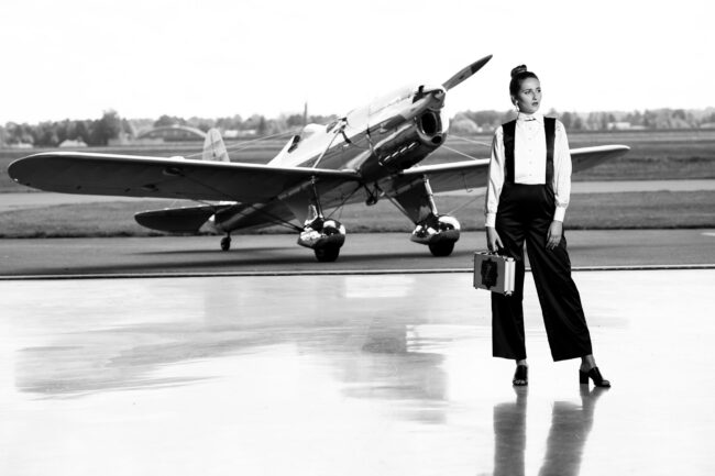 Fashion, Model in schwarz-weißem Hosenanzug und einem kleinen Koffer in der Hand vor Propellerflugzeug, schwarz-weiß