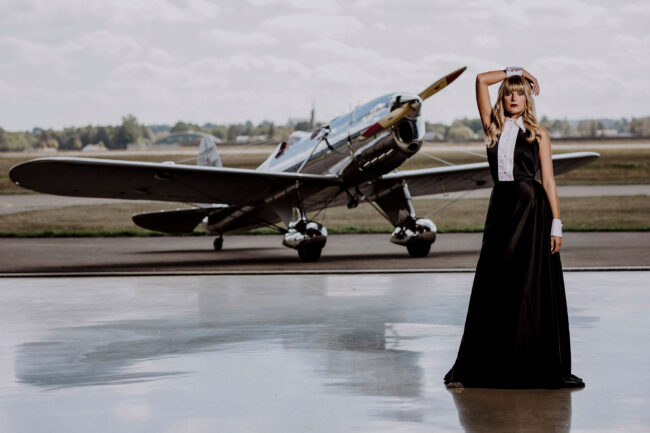 Fashion, Model schwarz weißem Kleid, ärmellos und weißen Manschetten vor Propellerflugzeug, Querformat