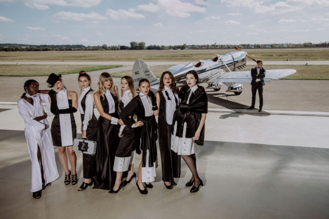 Fashion, Model Gruppen schwarz-weißen Kleidern vor Propellerflugzeug, im Hintergrund männliches Model in schwarzem Anzug und weißem Hemd bei Flugzeug stehend