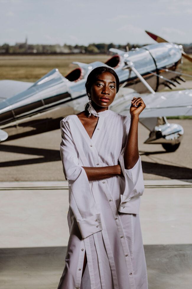 Fashion, Mode, dunkelhäutiges Model in weißen langen Kleid, vor silbernen Propellerflugzeug, Halbaufnahme