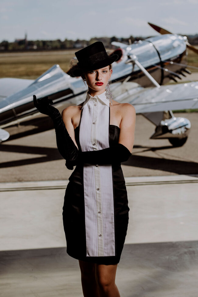 Fashion, Model mit schwarzem Hut, Handschuhen und weiß-schwarzes knielangen Kleid, vor Propellerflugzeug, Hochformat, Halbaufnahme