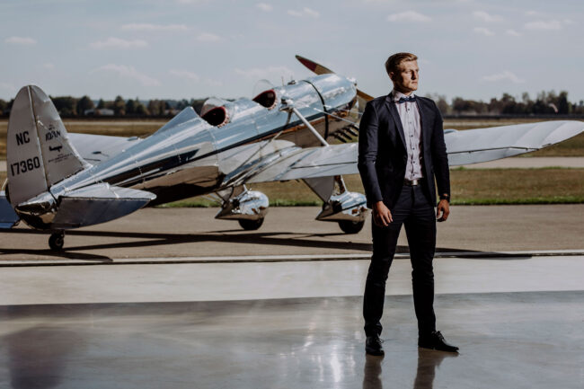 Fashion, männliches Model vor Propellerflugzeug, in schwarzem Anzug und weißem Hemd mit Fliege, Querformat