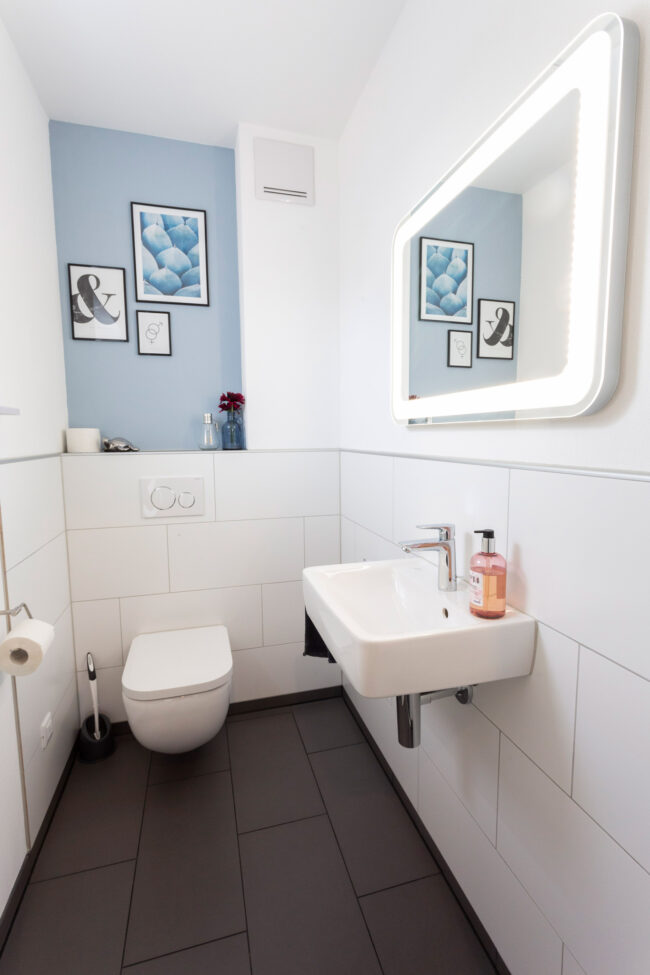 Architektur, Interieur Badezimmer mit schwarzen Bodenfliesen und weißen Kacheln an der Wand, mit Toilette, Waschbecken und beleuchtetem Spiegel