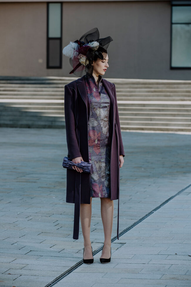 Fashion, Model in aubergine-farbenem Mantel und dunkelviolettem geblümten Kleid mit passender Handtasche und Kopfschmuck auf gepflastertem Platz stehend, Ganzaufnahme