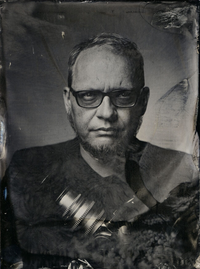 Nassplatte, Schwarz-Weiß, Portraitaufnahme von Mann mit Backenbart und schwarzer Brille, in der Hand hält er eine Kamera