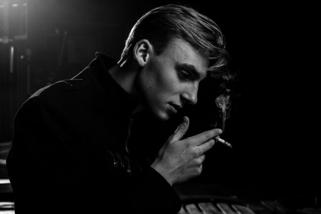 Fashion, Model, Portaritaufnahme eines jungen Mannes im Profil, hält Zigarette in der Hand, Hintergrund schwarz, schwarz-weiß