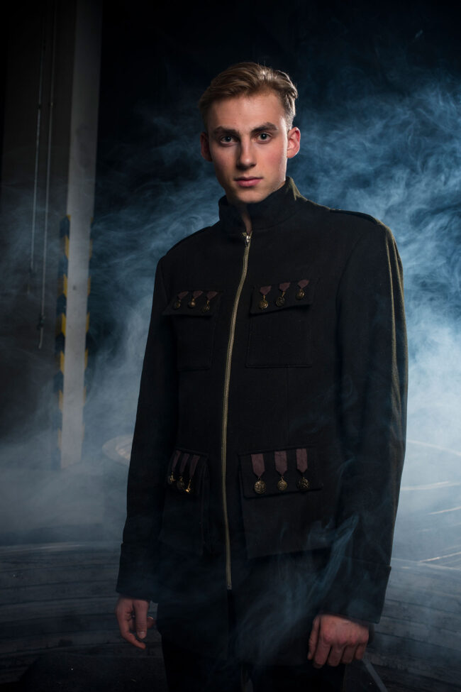 Fashion, Halbbildern von jungen Mann mit dunkelgrüner Jacke mit Reisverschluss. Auf der Jacke sind goldenen Orden ähnliche Verzierungen, im Hintergrund ist Nebel