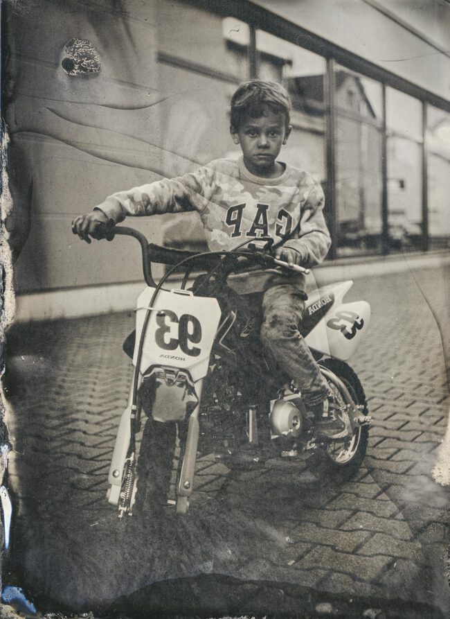 Nassplatte, Schwarz-Weiß, Ganzaufnahme von einem Jungen auf einer kleinen Motorkrossmaschine vor einem Gebäude