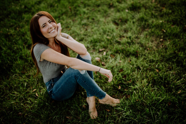 Portrait, Frau sitzt mit überkreuzten Beinen auf grüner Wiese und schaut lächelnd in Kamera hoch