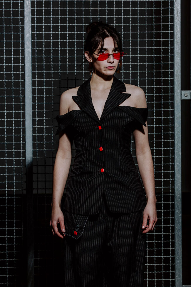 Fashion, Frau in schwarzem schulterfreien Nadelstreifenanzug mit roten Knöpfen und roter Sonnenbrille vor Metallgitter, Halbaufnahme, Hochformat