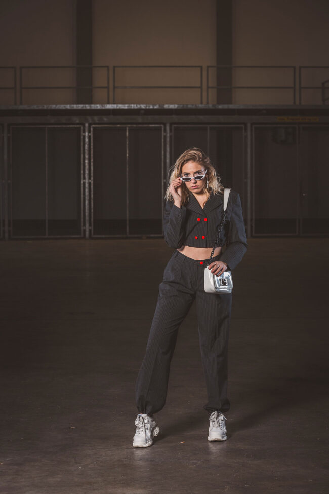 Fashion, Model mit schwarzer Hose und Bluse mit roten Knöpfen, Weiße Schuhe, Tasche und Sonnenbrille vor dunkler Halle